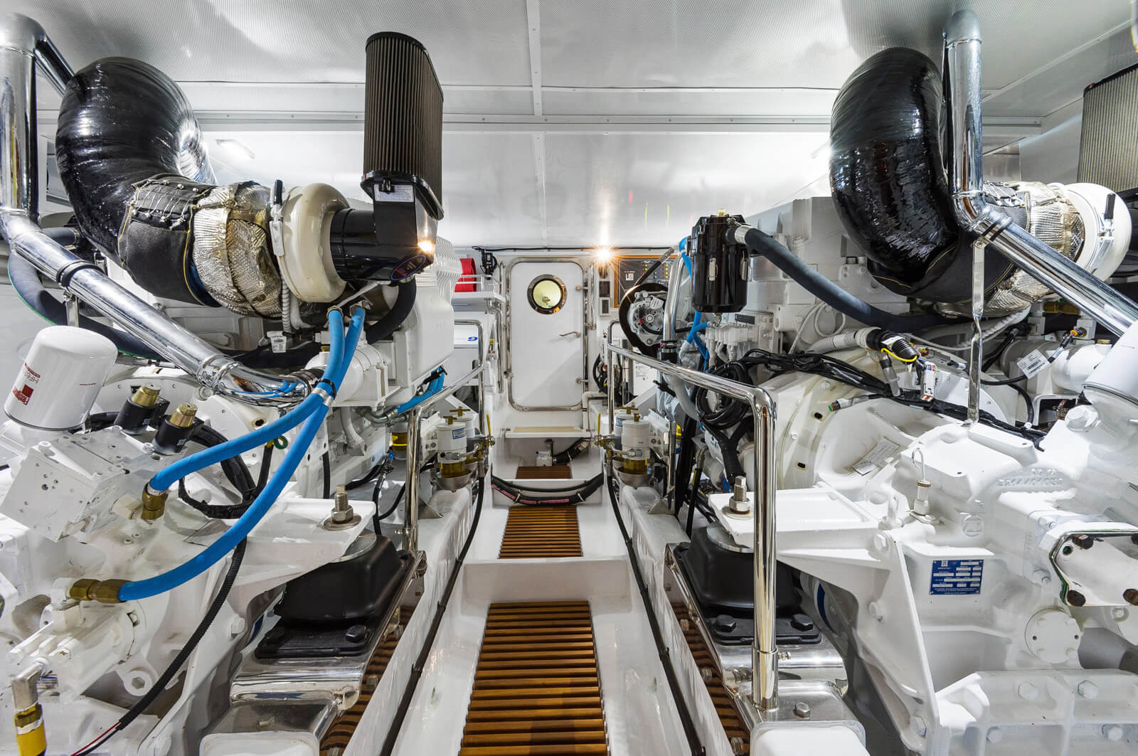 A clean, well-lit engine room housing two large, white marine engines with black and blue hoses, metallic pipes, and a grated walkway leading to a closed door at the far end.