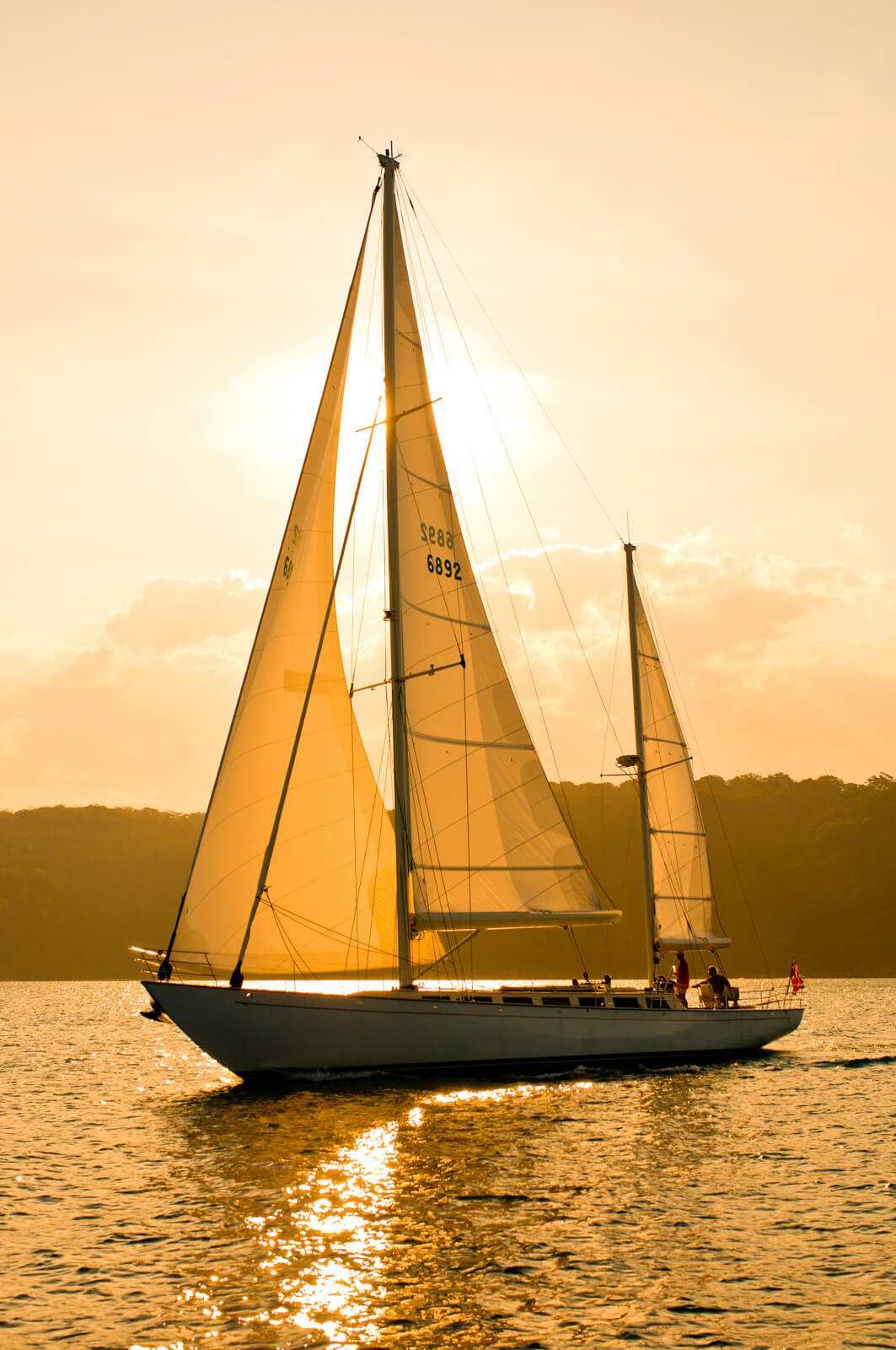 A sailboat with two sails glides on calm water at sunset, with golden sunlight illuminating the sails and reflecting off the water. There are people visible on the boat, and tree-covered hills are in the background.