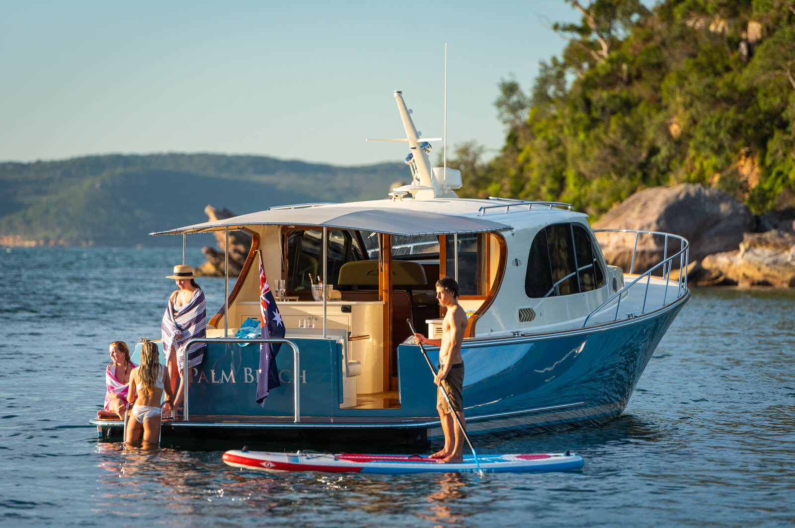 Four people relax near a blue and white yacht named Palm Beach, docked by a scenic, tree-lined shoreline. Two sit at the back, one stands with a paddleboard, and another is wrapped in a towel.