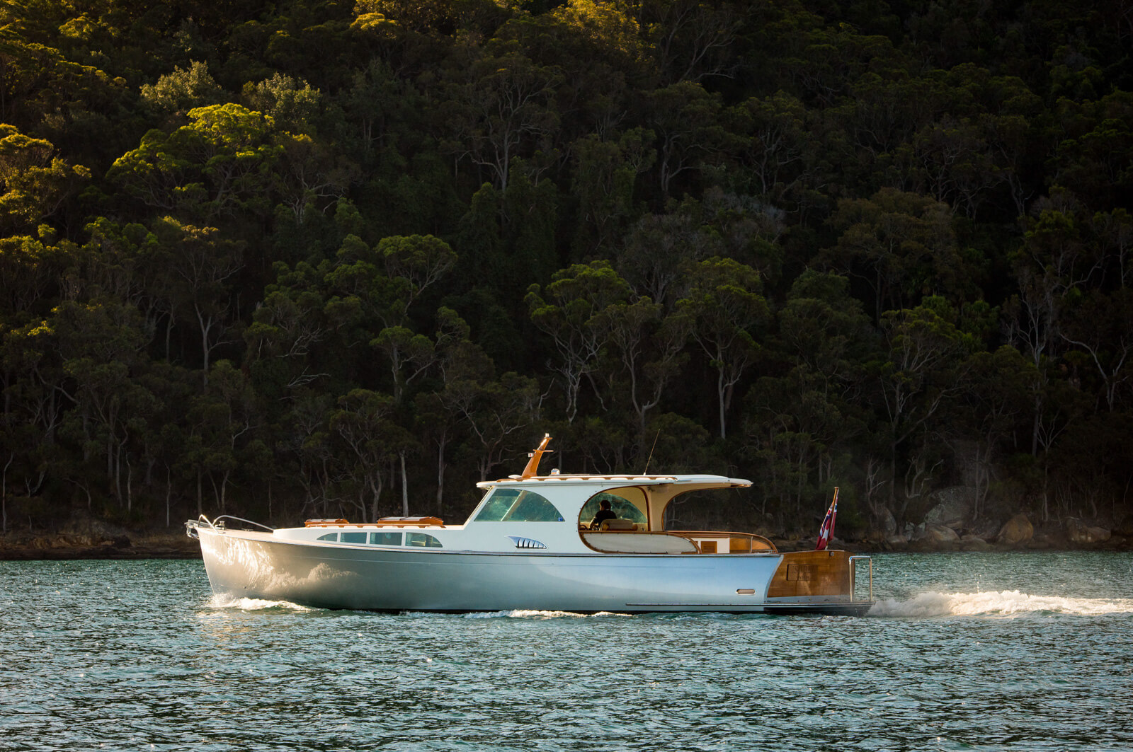 A sleek white motor yacht cruises on calm water near a forested shoreline, with tall green trees in the background and sunlight highlighting the boats polished wooden deck.