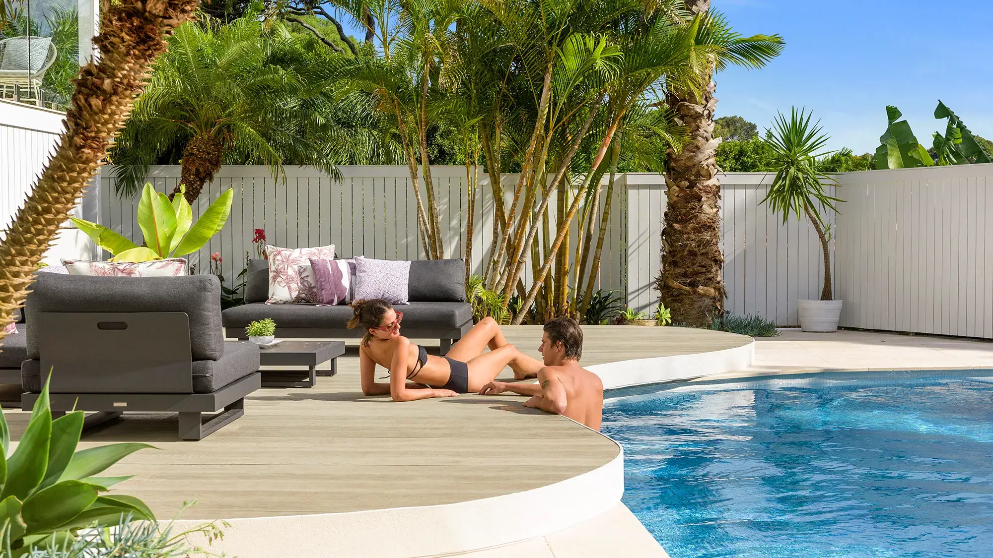 Two people relax by a pool surrounded by palm trees and tropical plants. One lies on a deck wearing sunglasses, the other is in the water. Outdoor furniture and bright sunshine create a summery, inviting atmosphere.