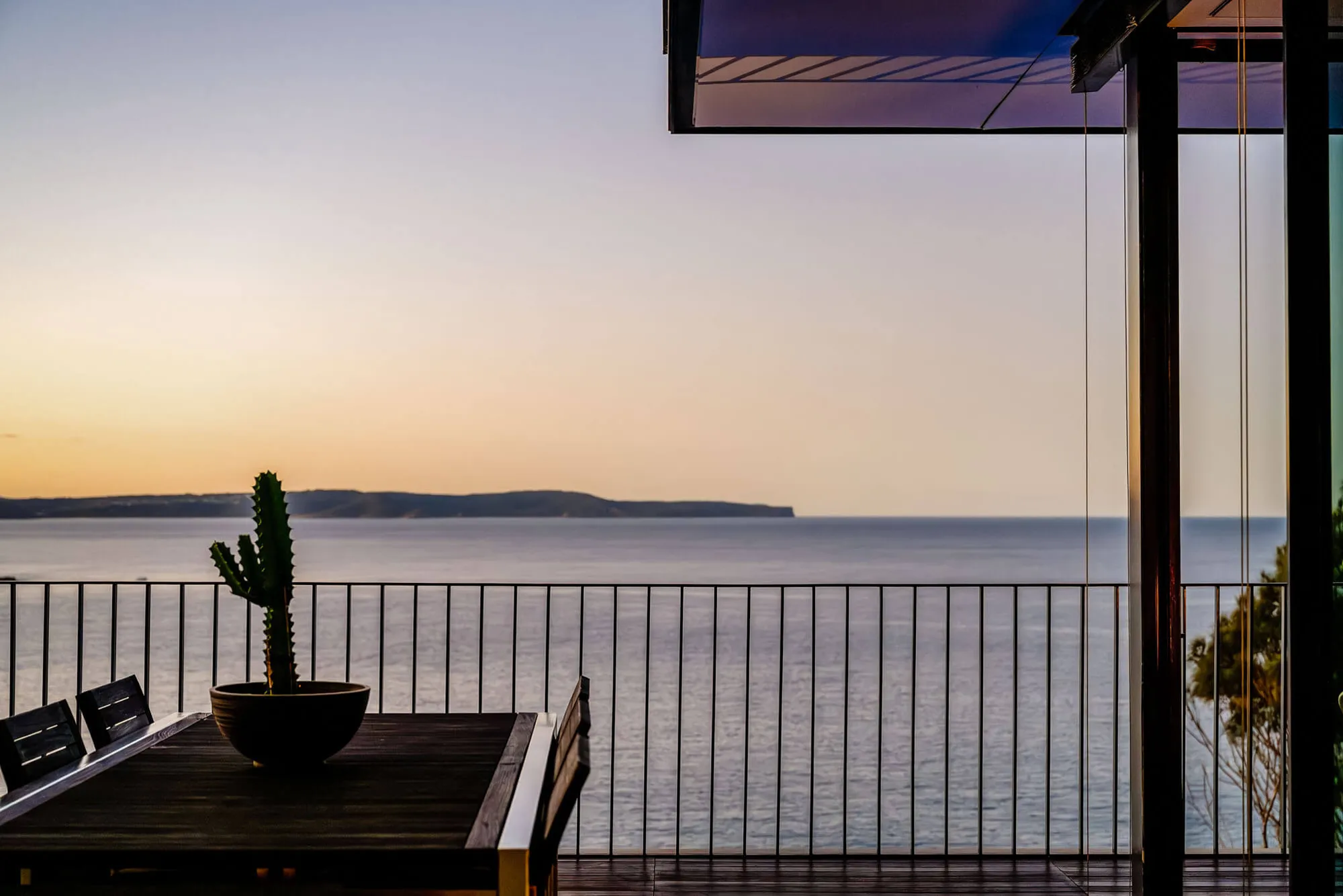 A balcony overlooking calm sea at sunset in Palm Beach, with a table, chairs, and a potted cactus in the foreground. A distant peninsula is visible across the water under a clear sky.