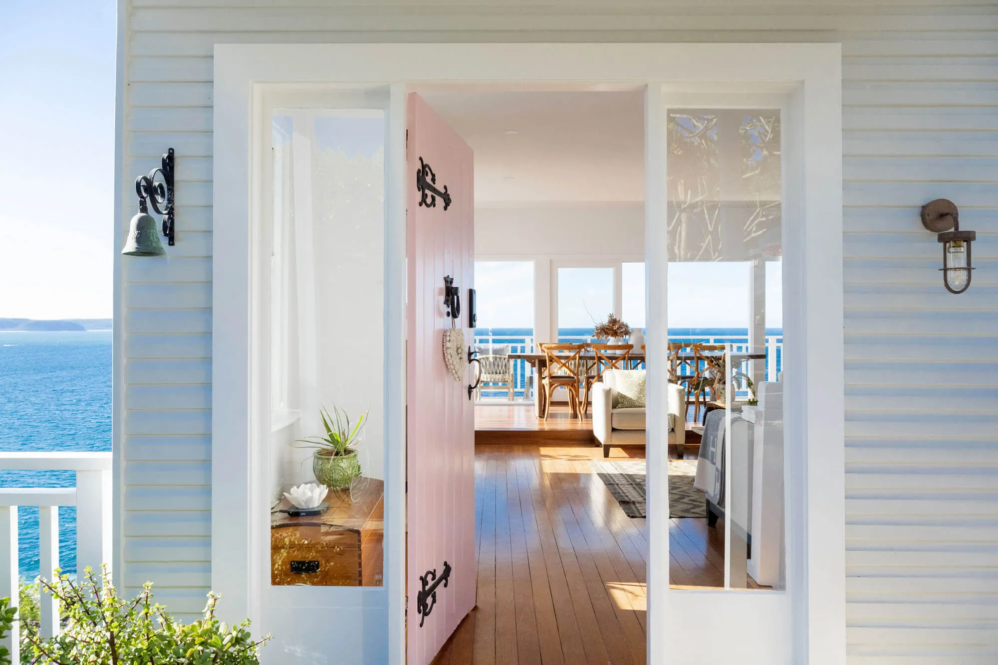 A pink front door opens to a bright, airy living space with wooden floors, white furniture, and large windows overlooking a blue ocean and sky. Plants and a decorative table are near the entrance.